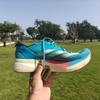 A hand holds up a pair of Adidas Adizero Adios Pro 3 running shoes in bright turquoise blue with white three-stripe logo, coral-orange midsole accents, and cream-colored sole, photographed outdoors in a park with green grass and trees under a clear blue sky