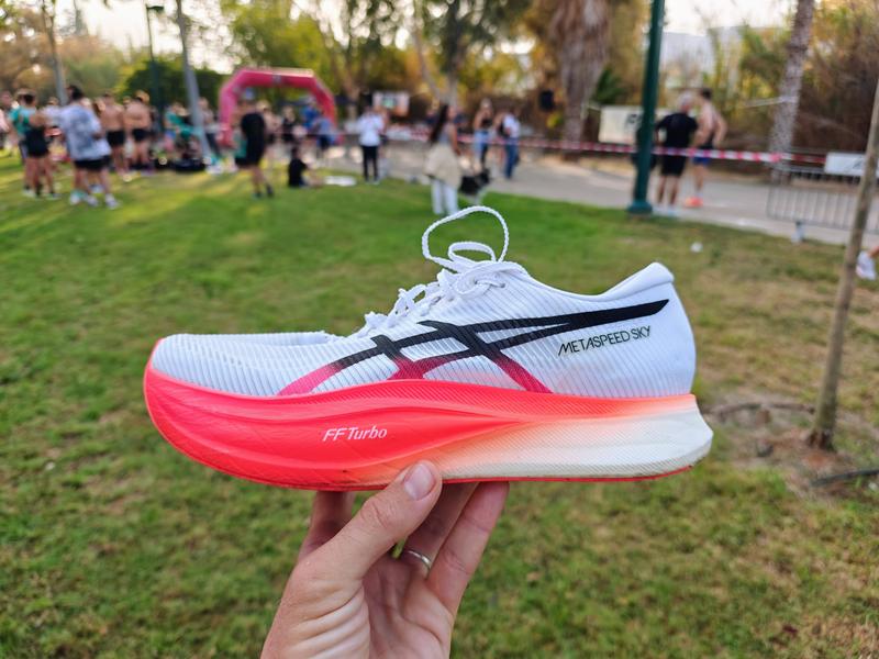 A hand holds up an ASICS Metaspeed Sky running shoe with white mesh upper, black ASICS stripes, and bright coral-pink FF Turbo midsole, photographed at a race event with a pink inflatable finish arch, crowd, and race barriers visible in the blurred background