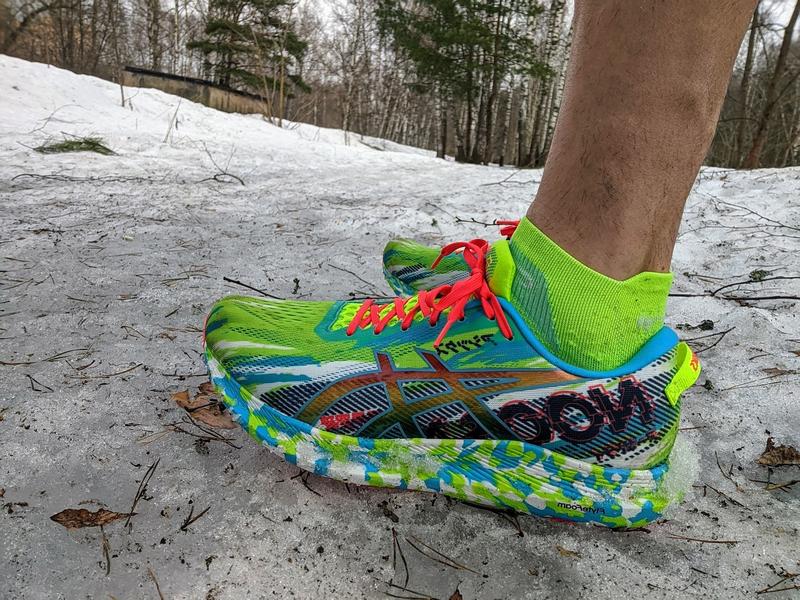 Close-up of my foot wearing a brightly colored ASICS Noosa Tri 13 running shoe in neon yellow-green with blue accents and red laces, standing on the ground with snow-covered forest terrain and birch trees visible in the background