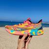 A hand holds up an ASICS Noosa Tri running shoe in vibrant neon pink, yellow, and turquoise with colorful geometric patterns and a multi-colored striped sole featuring FlyteFoam technology, photographed on a sunny beach with blue sea and clear sky in the background