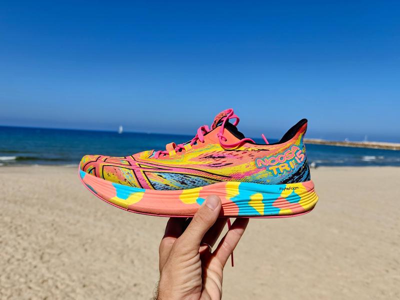 A hand holds up an ASICS Noosa Tri running shoe in vibrant neon pink, yellow, and turquoise with colorful geometric patterns and a multi-colored striped sole featuring FlyteFoam technology, photographed on a sunny beach with blue sea and clear sky in the background