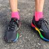 Overhead view of a runner's feet wearing black Mizuno running shoes with rainbow gradient accents (red, yellow, green, blue) on the sole and bright pink ankle socks, standing on gray asphalt pavement