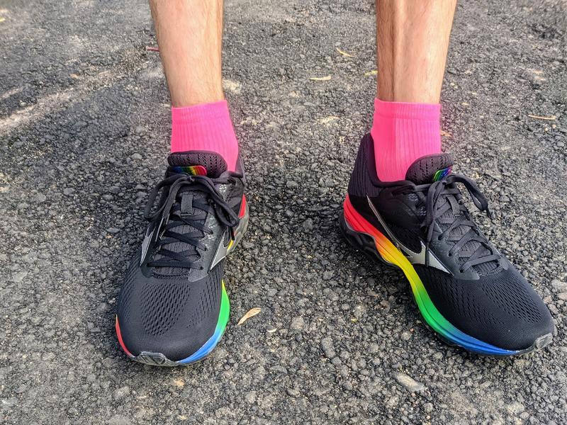 Overhead view of a runner's feet wearing black Mizuno running shoes with rainbow gradient accents (red, yellow, green, blue) on the sole and bright pink ankle socks, standing on gray asphalt pavement