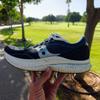A hand holds up a well-worn Saucony running shoe with navy blue and gray upper and a dirty, cracked white sole, photographed outdoors in a sunny park with green lawns, trees, and a paved path visible in the background