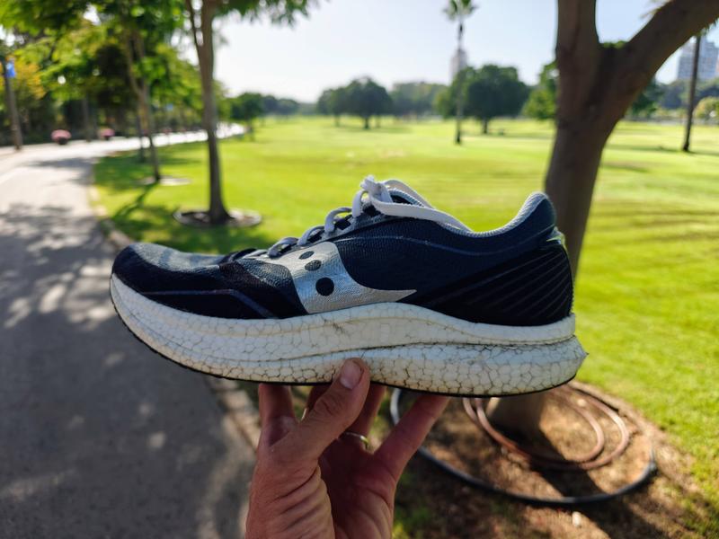 A hand holds up a well-worn Saucony running shoe with navy blue and gray upper and a dirty, cracked white sole, photographed outdoors in a sunny park with green lawns, trees, and a paved path visible in the background