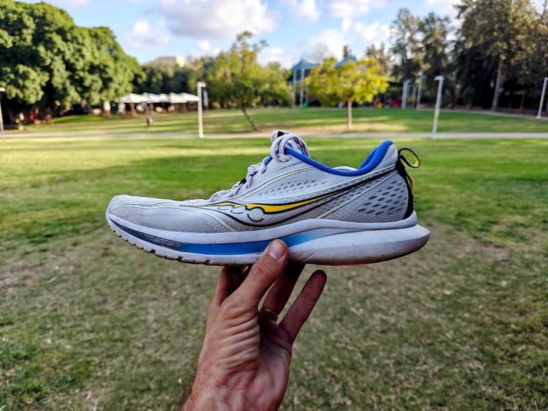 A hand holds up a Saucony Kinvara 13 running shoe in white color with blue accents, yellow and black stripe logo, and white sole, photographed outdoors in a sunny park with green lawn and trees.