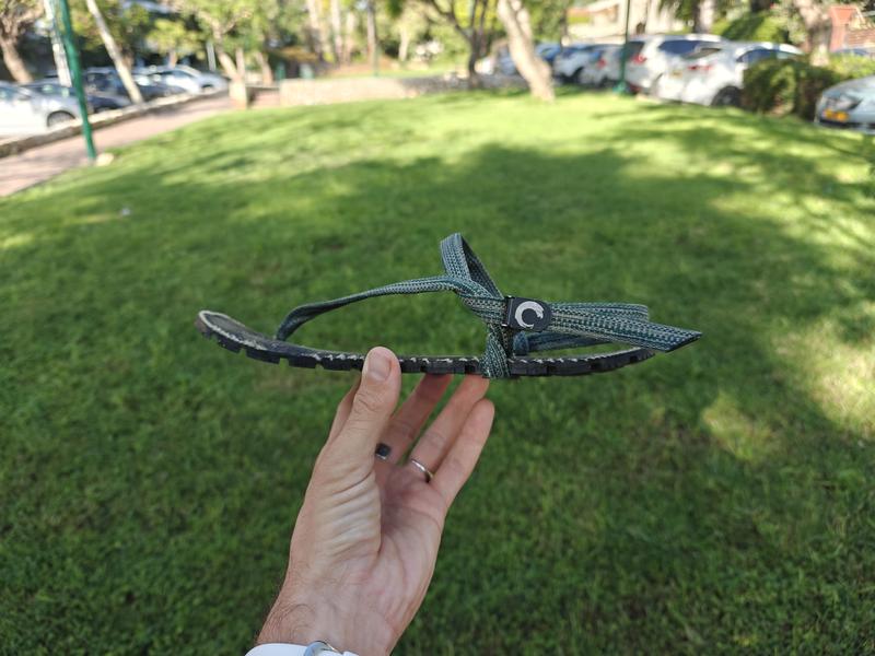 Overhead view of my feet wearing dark green strapped Zuzu Flow sandals standing on the ground scattered with pine cones, dry grass, and pine needles