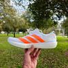 A white Adidas Adios 9 running shoe with bright orange stripes is held in a hand in the foreground. The shoe is shown in side profile, with white laces and a thick, light-colored midsole. The background is an outdoor park scene with green grass and leafy trees, softly blurred