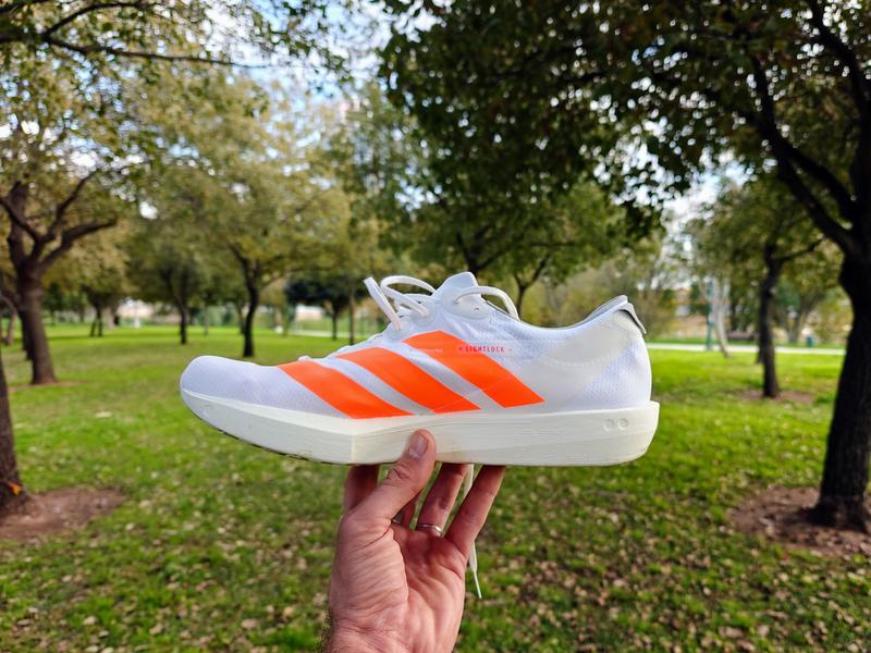 A white Adidas Adios 9 running shoe with bright orange stripes is held in a hand in the foreground. The shoe is shown in side profile, with white laces and a thick, light-colored midsole. The background is an outdoor park scene with green grass and leafy trees, softly blurred
