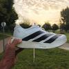 A hand holds up a white Adidas Adizero Evo SL running shoe against a soft, sunlit park background. The shoe features bold, diagonal black stripes and a thick, white Lightstrike Pro foam midsole. The setting is a paved path lined with green grass and trees during a hazy sunset.