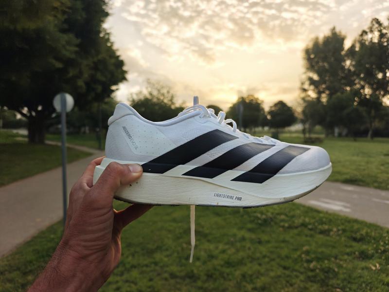 A hand holds up a white Adidas Adizero Evo SL running shoe against a soft, sunlit park background. The shoe features bold, diagonal black stripes and a thick, white Lightstrike Pro foam midsole. The setting is a paved path lined with green grass and trees during a hazy sunset.