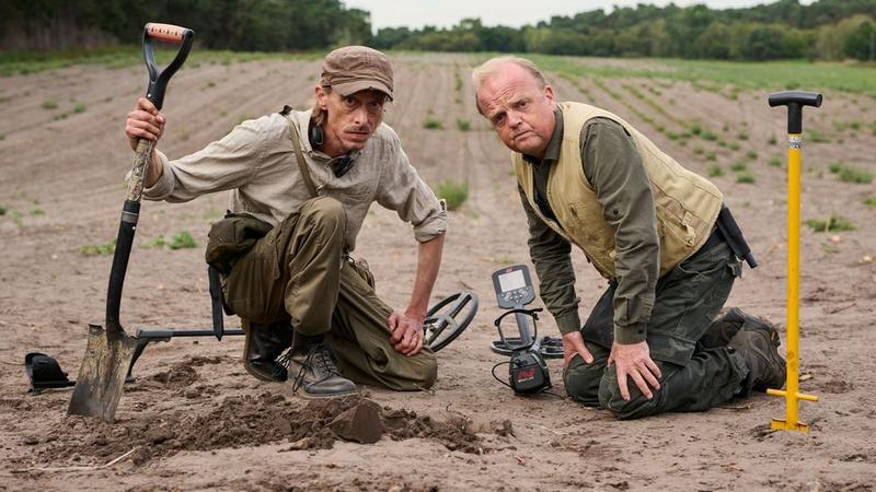 Two men kneel in a plowed field using metal detectors and shovels, examining a small hole in the ground, with countryside trees in the background; a scene from the TV show Detectorists