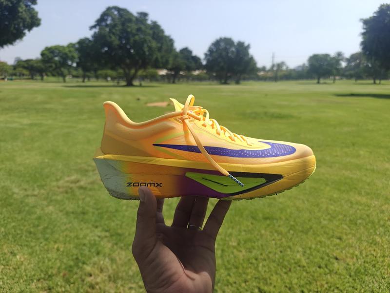 Yellow-orange Nike Zoom Fly 6 running shoe held up against a sunny green park landscape.