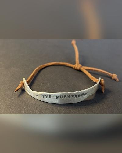 A curved silver bar stamped with the Russian words “и так нормально” (“good enough”), tied on a brown leather cord, on a dark grey surface.