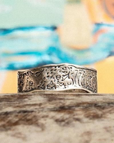 A wide band of oxidised silver covered in tiny engraved figures and symbols, on weathered wood against a painted backdrop.