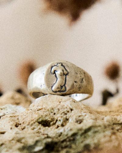 A chunky recycled-silver ring with a small engraved standing figure, resting on a rough stone.