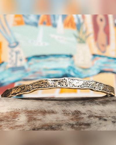 A thin hammered silver bangle engraved with sea motifs, lying on weathered wood in front of a colourful painting.